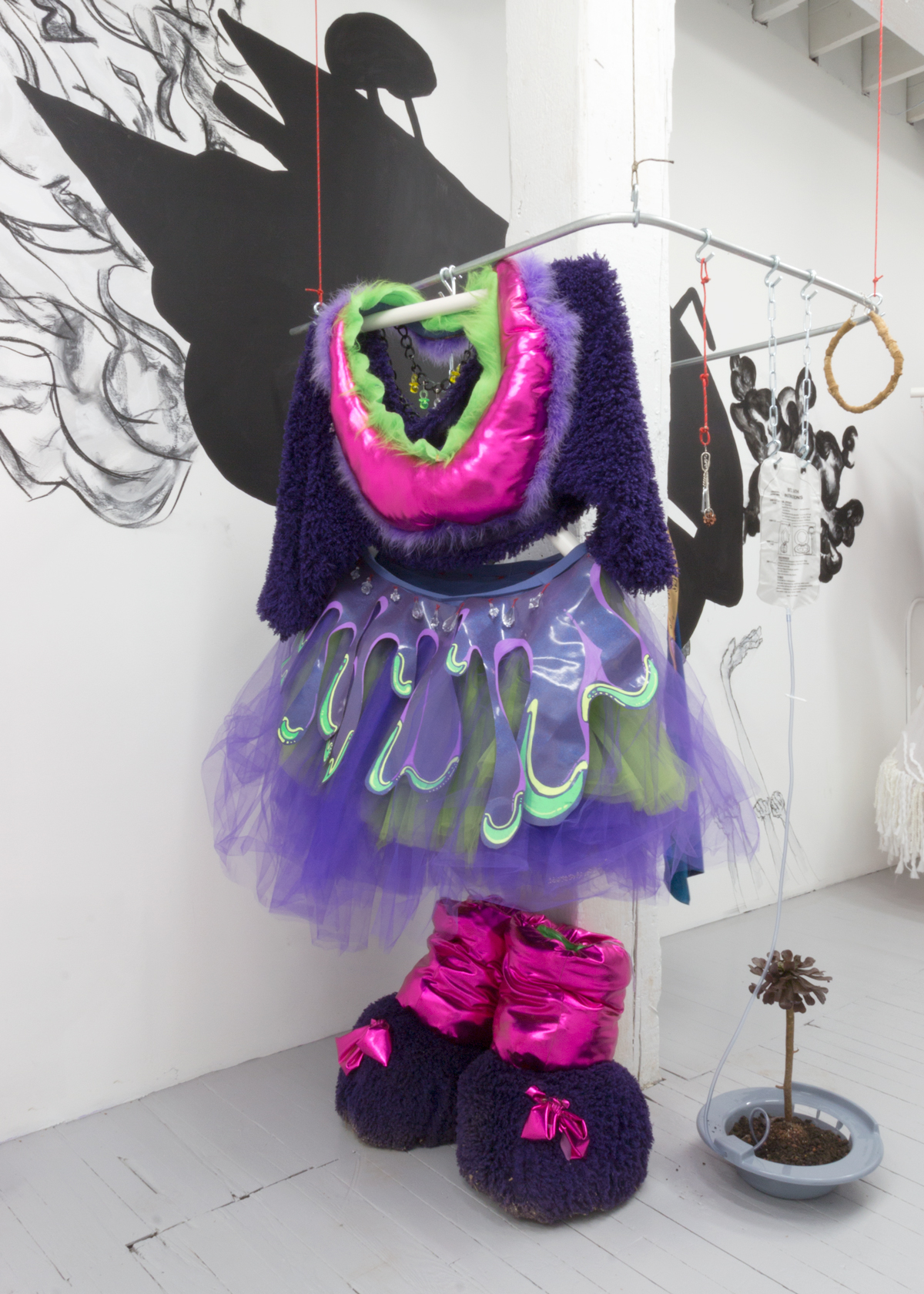 A colorful pink and purple costume hung on a metal rack with a sculpture of a plant in a sitz bath on the floor beside it